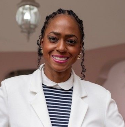 Smiling person wearing a white blazer over a blue and white striped shirt with a collar, standing indoors.