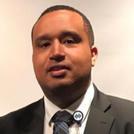 A person in a dark suit, white collared shirt, and patterned tie with an identification badge clipped to their collar.
