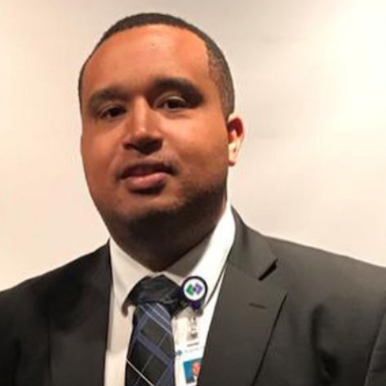 A person in a dark suit, white collared shirt, and patterned blue tie wearing a professional identification badge.