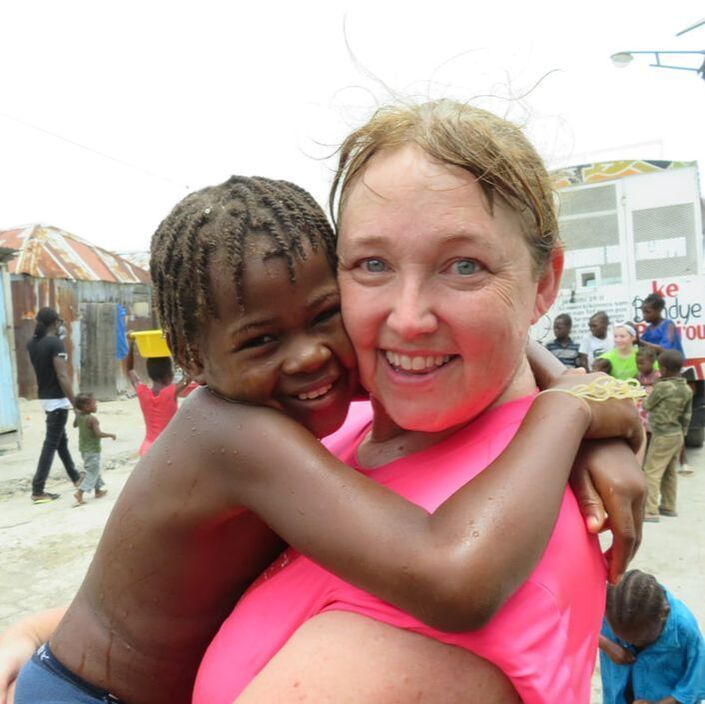 A smiling woman in a bright pink top is being hugged by a child in an outdoor setting with buildings and other people.