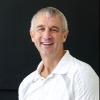 A smiling person with short, light hair wearing a white polo shirt against a black background.