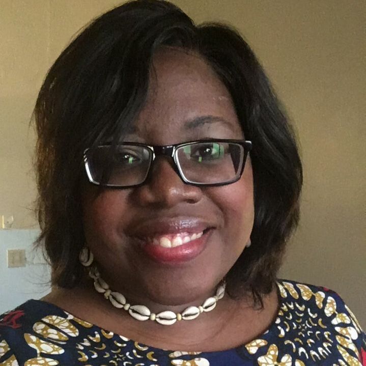A smiling person wearing black-rimmed glasses and a necklace made of cowrie shells.