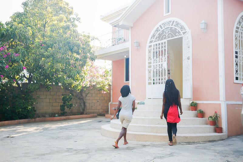 Two people walk toward the arched entrance of a peach-colored building with steps and potted plants.