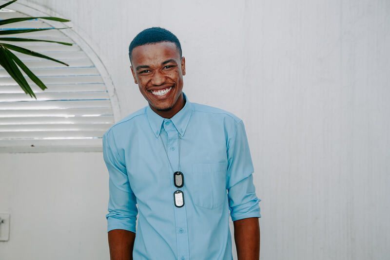 A smiling person wearing a light blue button-down shirt and dog tag necklace, standing in front of a white wall.