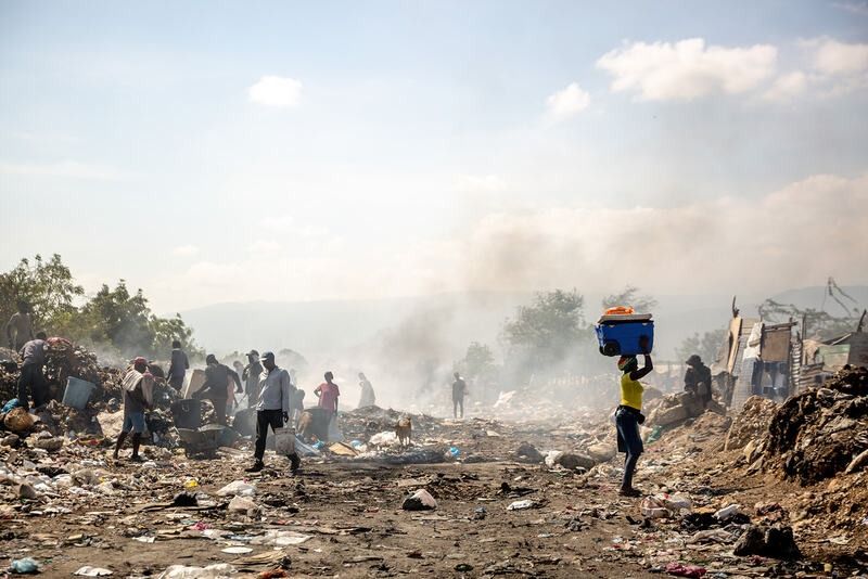 People walk through a smoky landfill, searching through piles of debris under a bright, hazy sky.