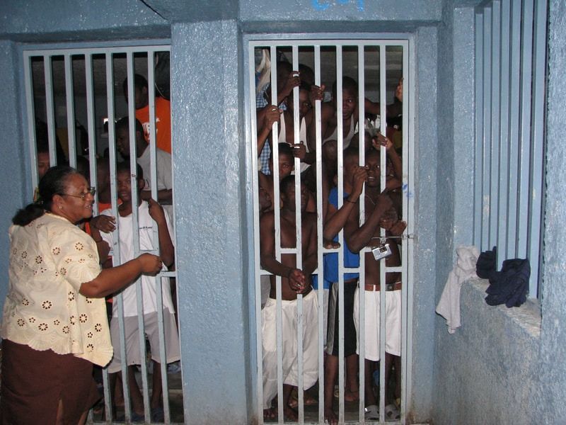 A woman stands outside a jail cell with barred doors, where several people are crowded inside looking out.