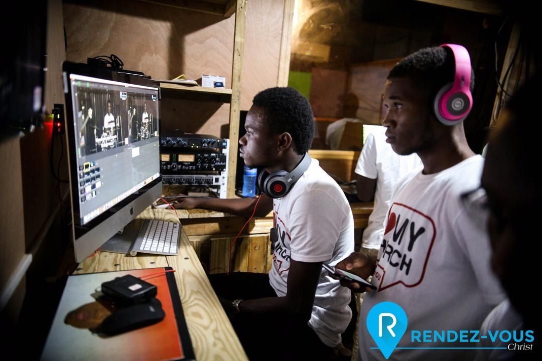 Two people in white t-shirts working at a computer in a dimly lit studio, one wearing pink headphones.