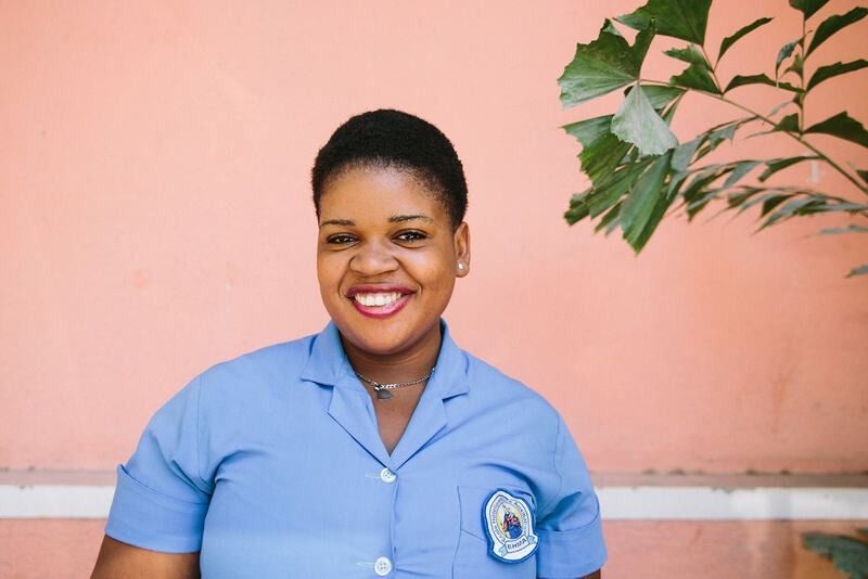 A person wearing a light blue uniform with a badge, smiling against a plain peach-colored wall with green leaves nearby.