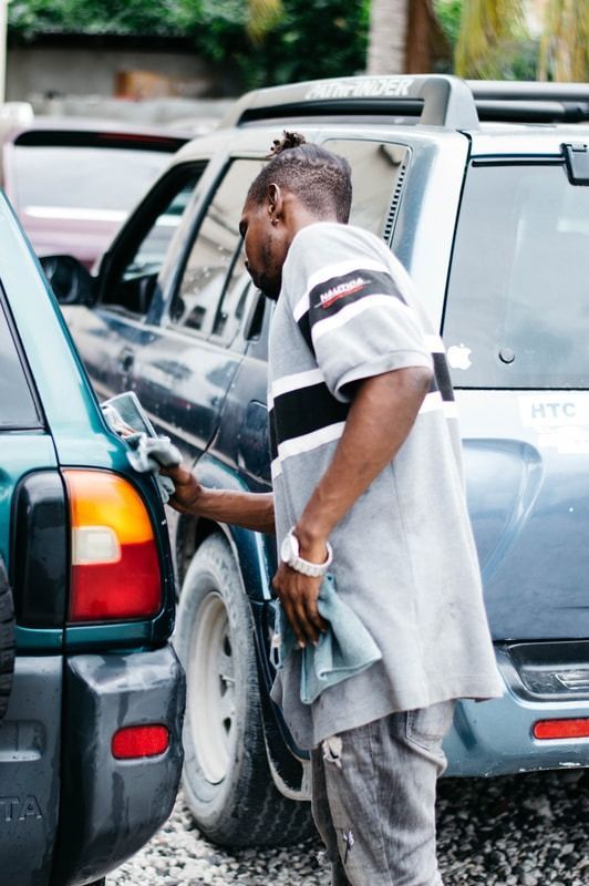A person wearing a grey and white shirt cleans the exterior of a dark green SUV in a gravel lot.