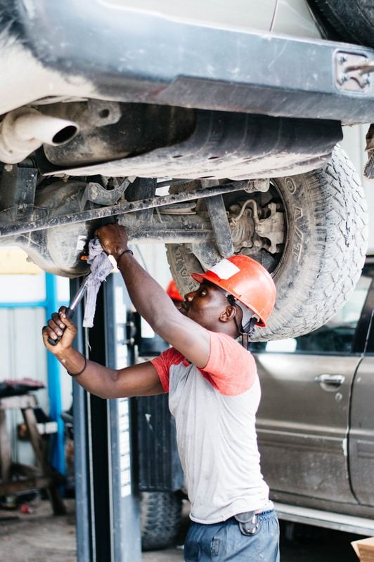 A mechanic wearing a hard hat uses a wrench to work on the underside of a lifted vehicle in a garage.