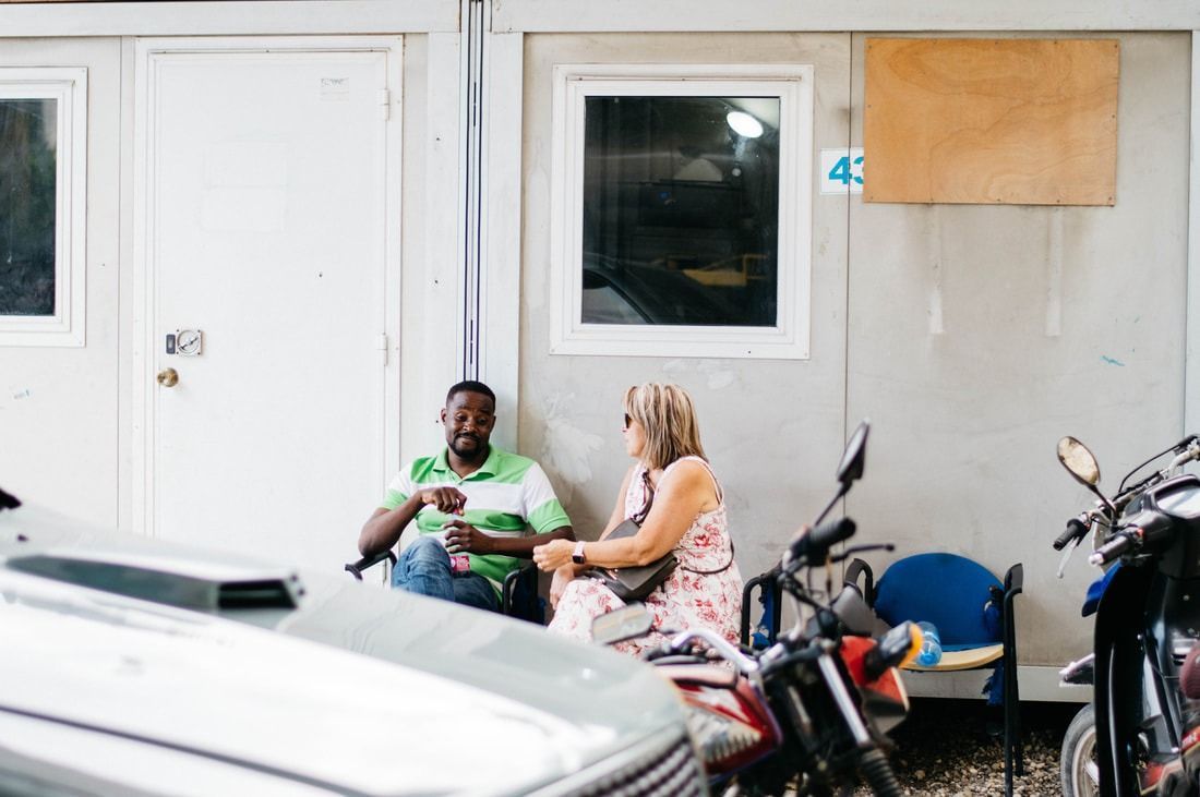 Two people sit on chairs outside a building, talking in a parking area near a motorcycle and a blue chair.