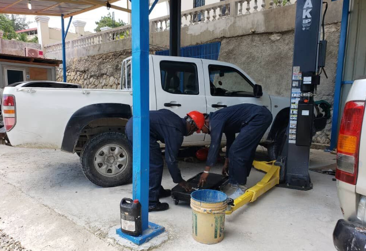 Two workers in blue uniforms and hard hats maintain a white pickup truck lifted on a hydraulic rack in a garage.