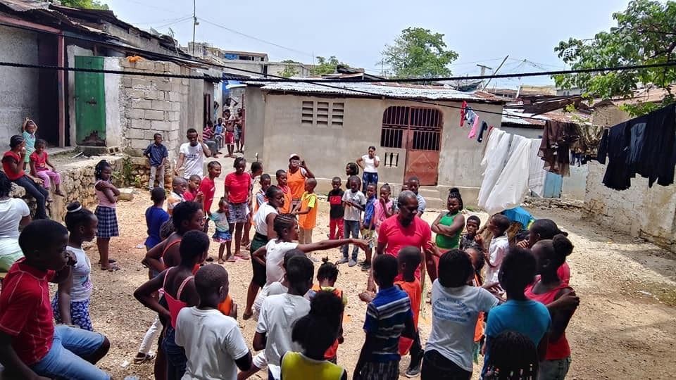 A diverse group of people gathers in a residential courtyard, engaging in a communal activity near small houses.