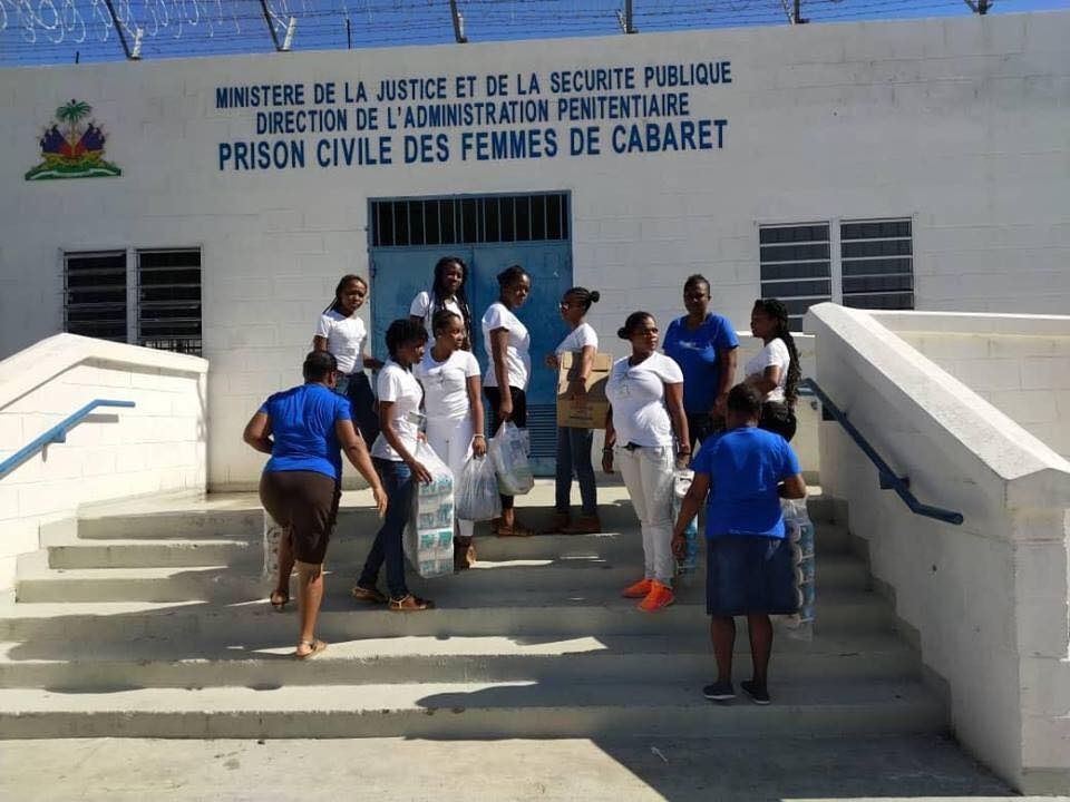 People stand on the steps of the Prison Civile des Femmes de Cabaret in Haiti, some carrying bags of supplies.