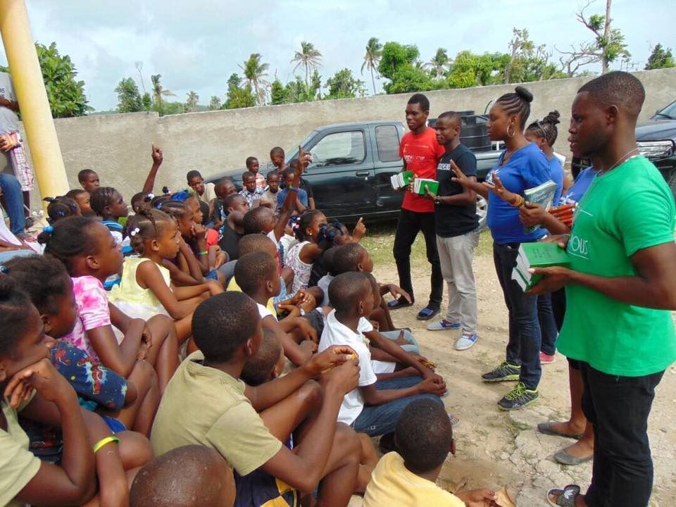 A group of people are outdoors distributing books to a large gathering of children seated on the ground.