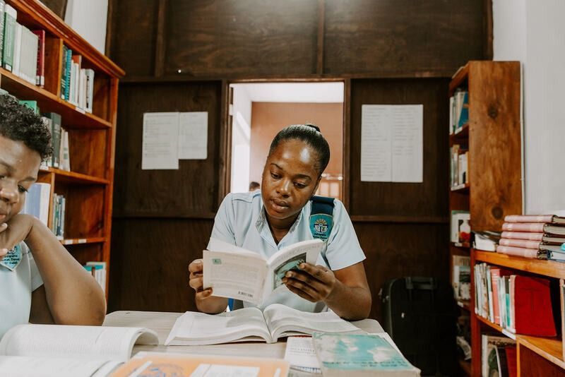 Two students wearing matching light blue uniforms sit at a table in a library, studying with open books.