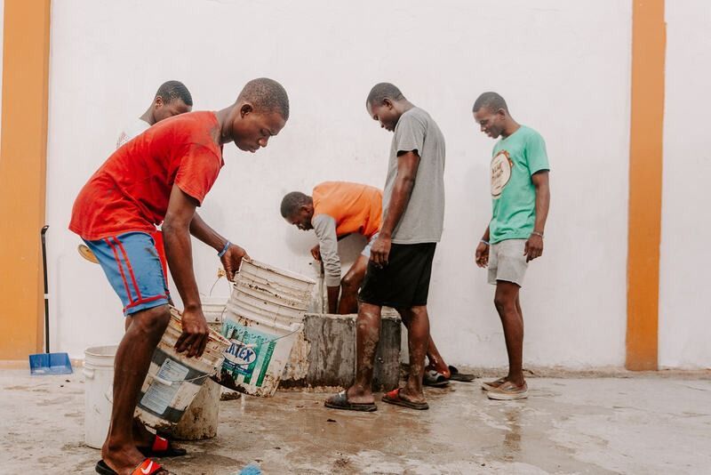 Five people work together to mix cement in buckets on a patio against a white wall.