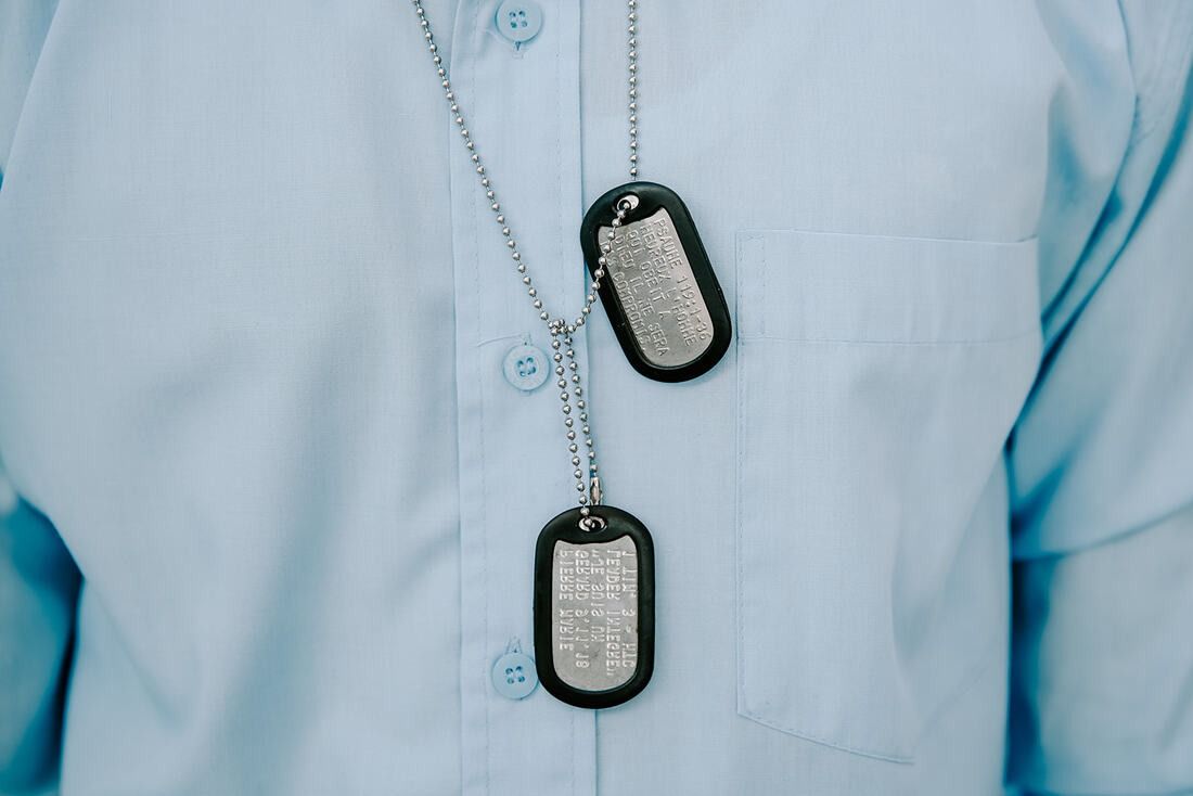 A person wearing a light blue button-down shirt with two silver military-style dog tags hanging on a ball chain.