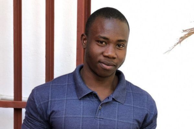 A person wearing a blue checkered polo shirt, looking at the camera with a neutral expression in front of a window grate.