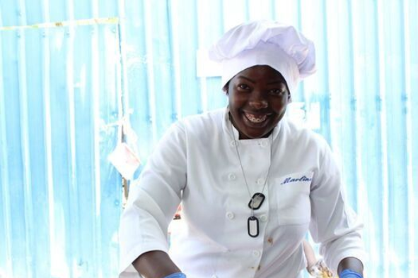 A smiling chef in a white uniform and hat works in a kitchen, wearing blue gloves and glasses around their neck.