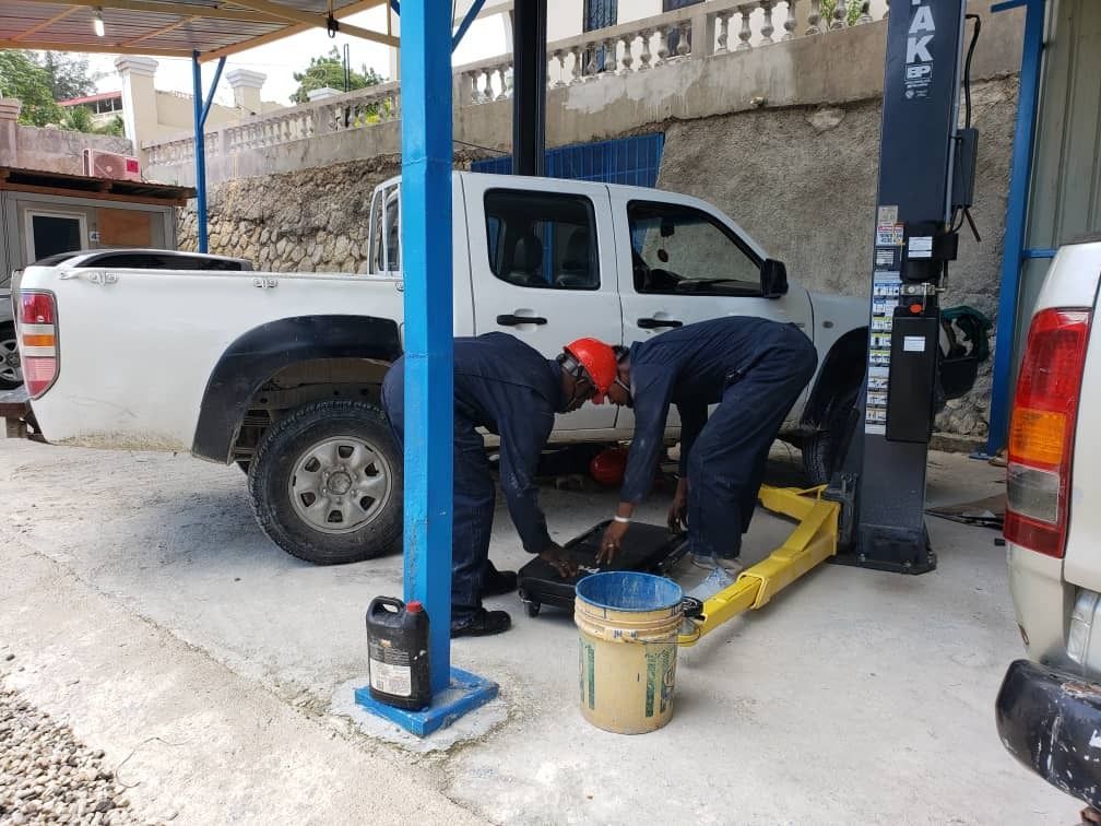 Two mechanics in dark uniforms working under a white pickup truck lifted on a hydraulic garage hoist.