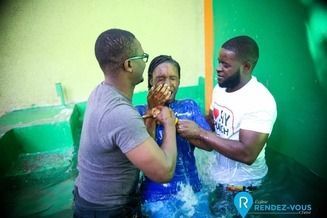 Two people assist a woman during a religious water baptism ceremony in a pool with bright green walls.