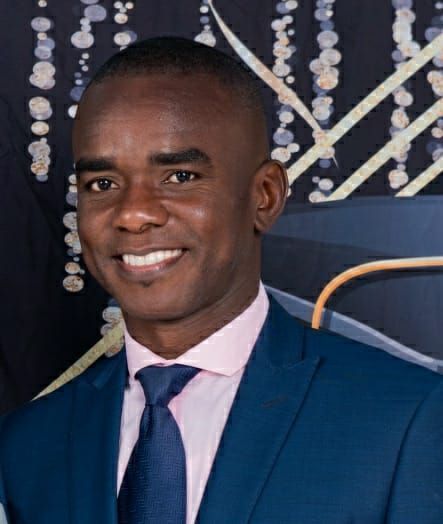 A person smiling while wearing a blue suit, light pink collared shirt, and blue tie in front of a decorative backdrop.