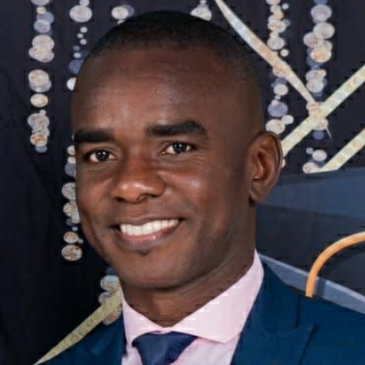 A smiling person wearing a dark blue suit, light pink shirt, and dark blue tie against a dark, decorative background.