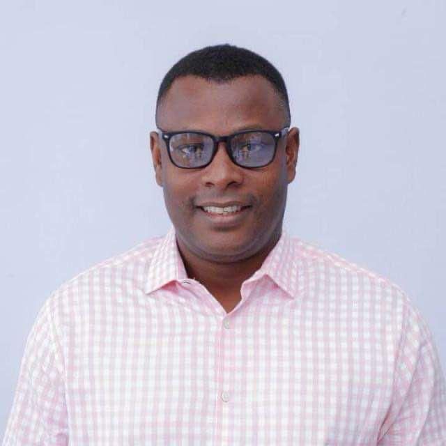 A person wearing black-rimmed glasses and a light pink gingham button-down shirt, smiling against a plain white background.