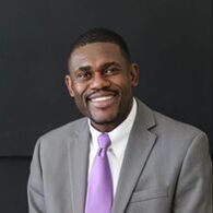 A smiling person wearing a grey suit, white collared shirt, and purple tie against a dark background.