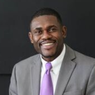 A smiling person wearing a grey suit, white collared shirt, and purple tie against a plain black background.