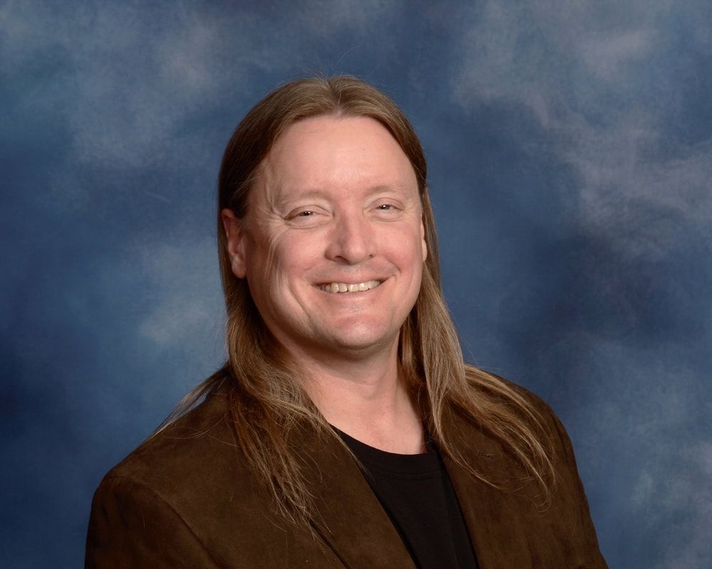 A person with long hair wearing a brown blazer, smiling in front of a blue, cloudy backdrop.