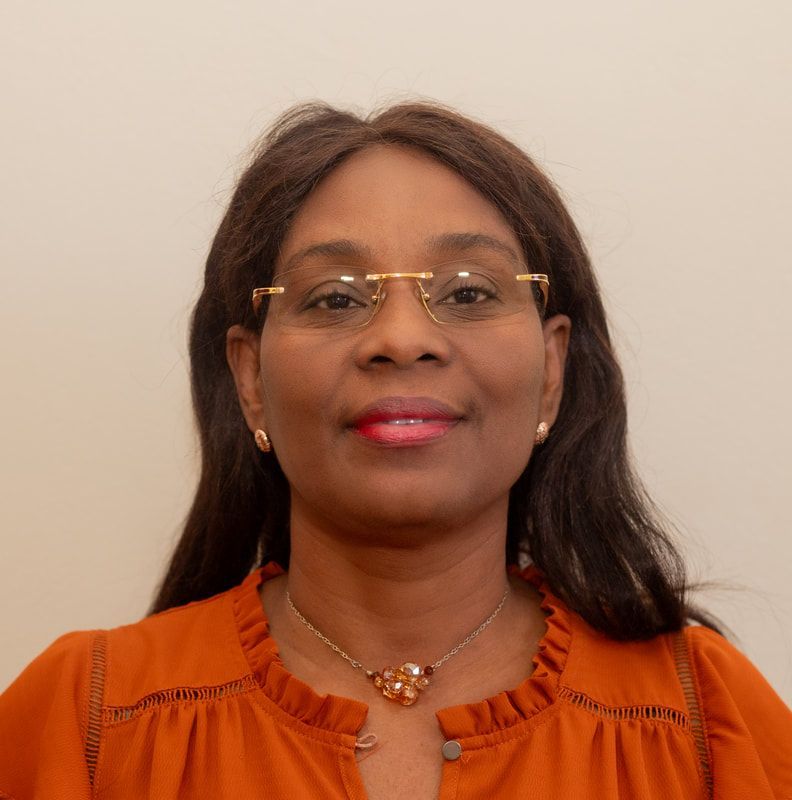 A person with dark hair and glasses wears a rust-colored top and a statement necklace against a plain, light background.
