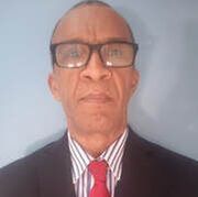 A person wearing glasses, a dark blazer, a striped button-down shirt, and a red tie, posed against a light blue background.