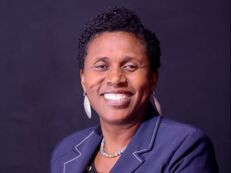 A smiling professional wearing a blue blazer and statement earrings against a dark background.