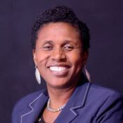 A person with short, dark, textured hair wears a navy blue blazer and large silver earrings, smiling against a dark background.