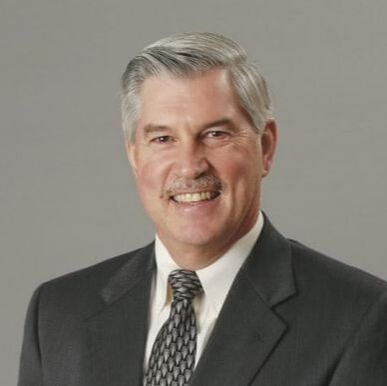 A smiling person wearing a dark grey suit and a patterned tie, posed against a solid grey background.