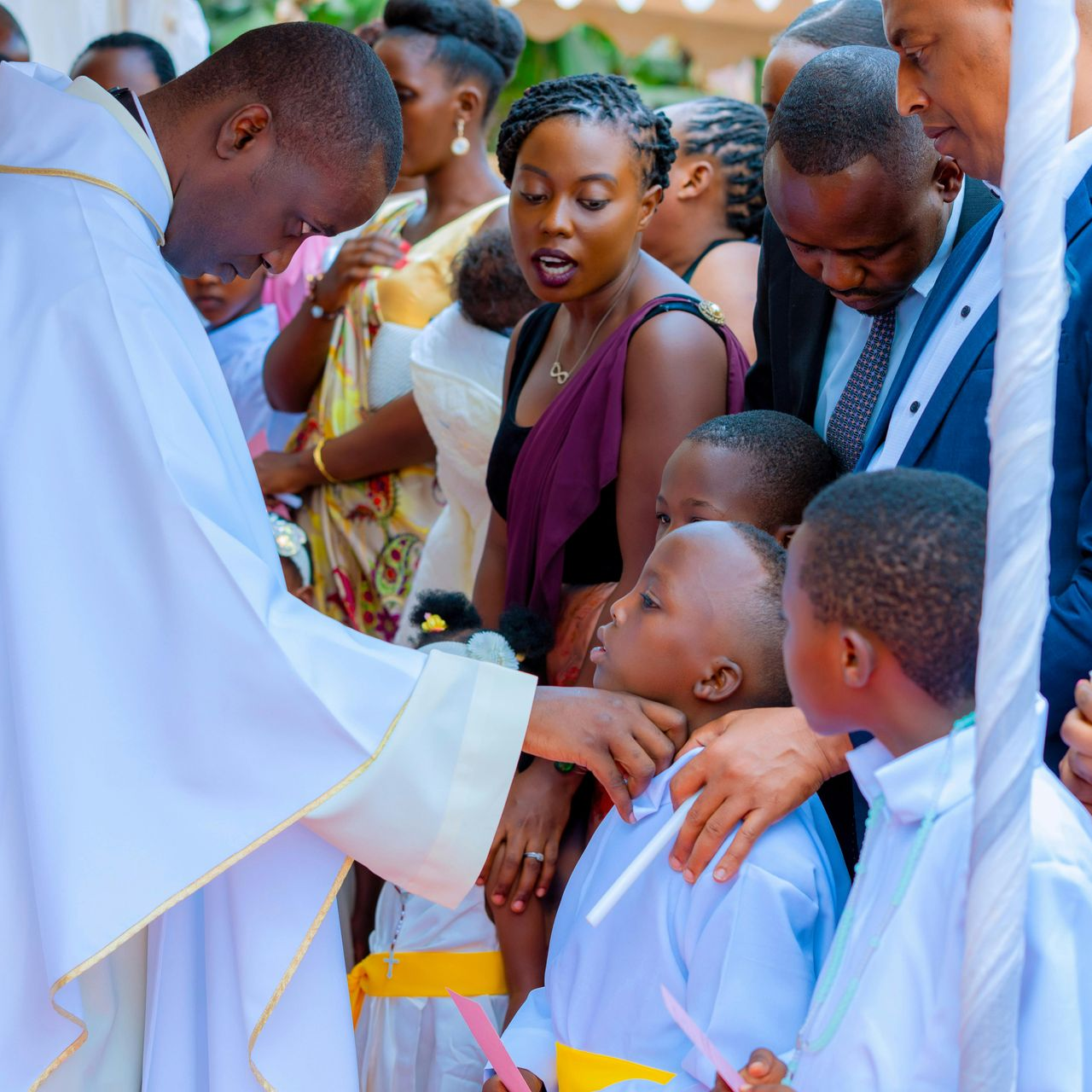 A priest in white vestments touches the forehead of a child during a religious ceremony with attendees nearby.