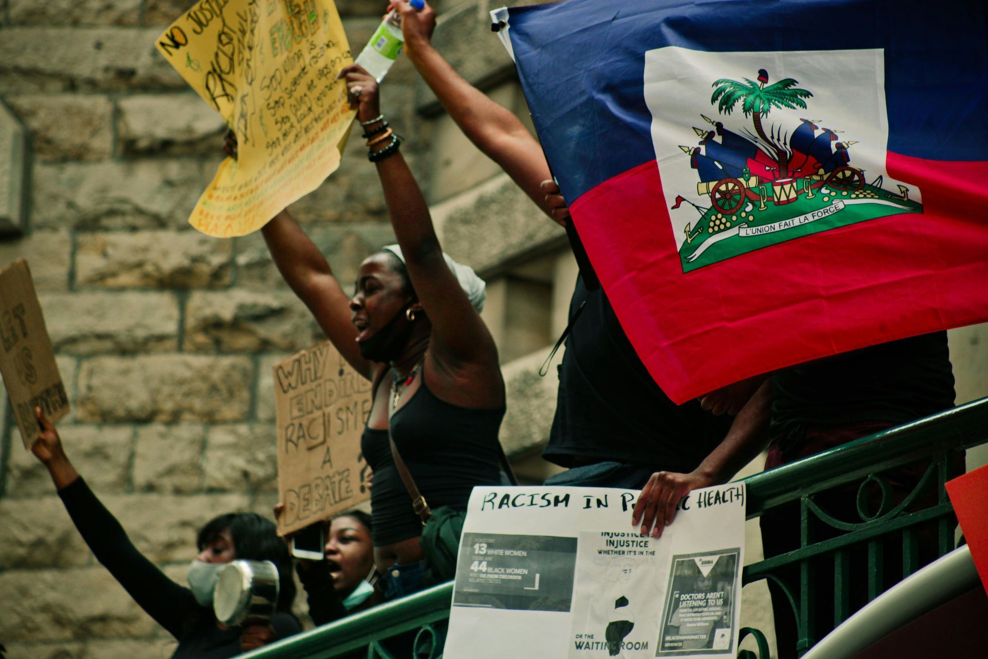 Protesters hold signs and a Haitian flag during a rally in front of a stone building.