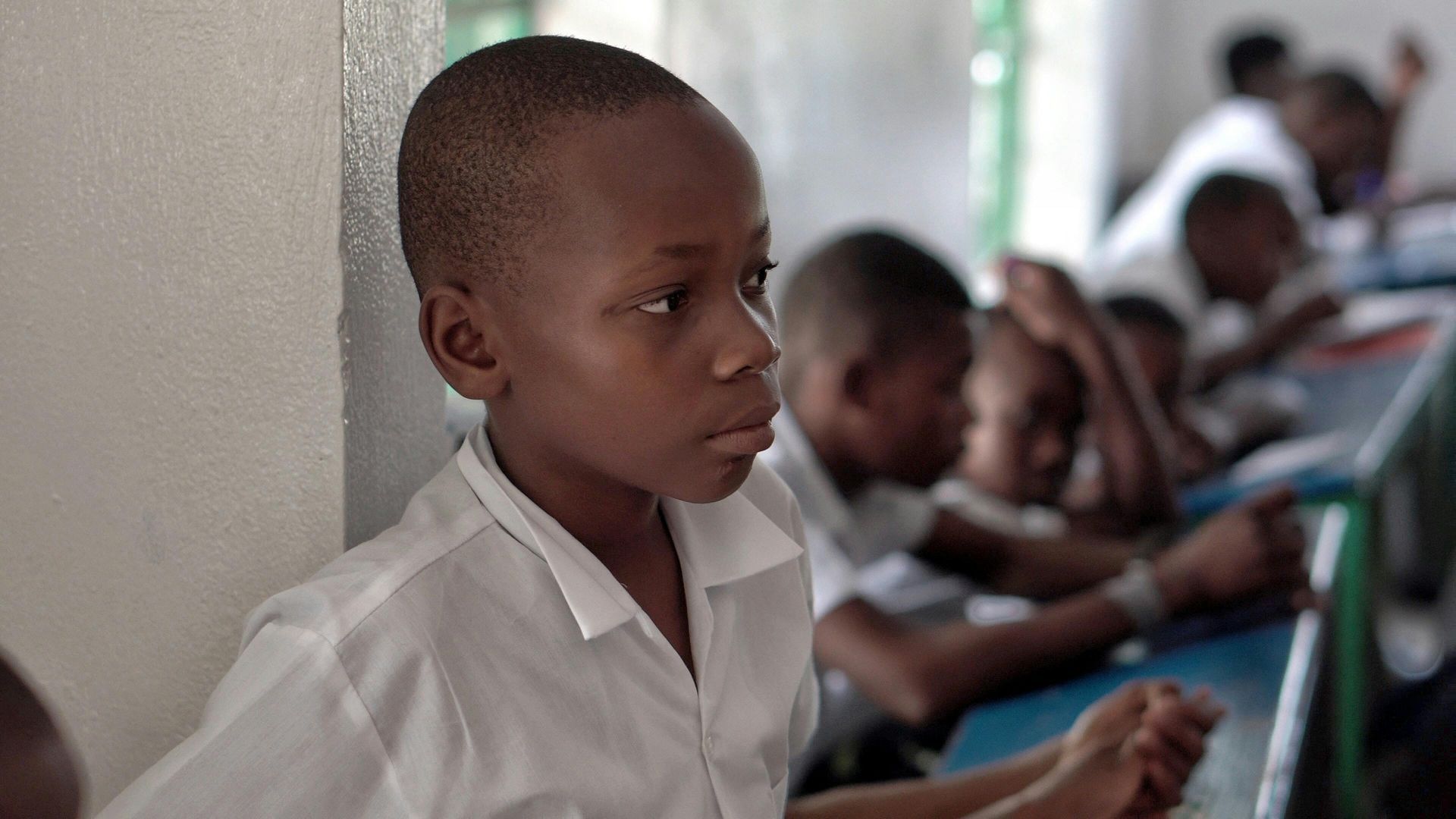 Students in white shirts sitting at desks in a classroom, with one student in the foreground looking to the side.