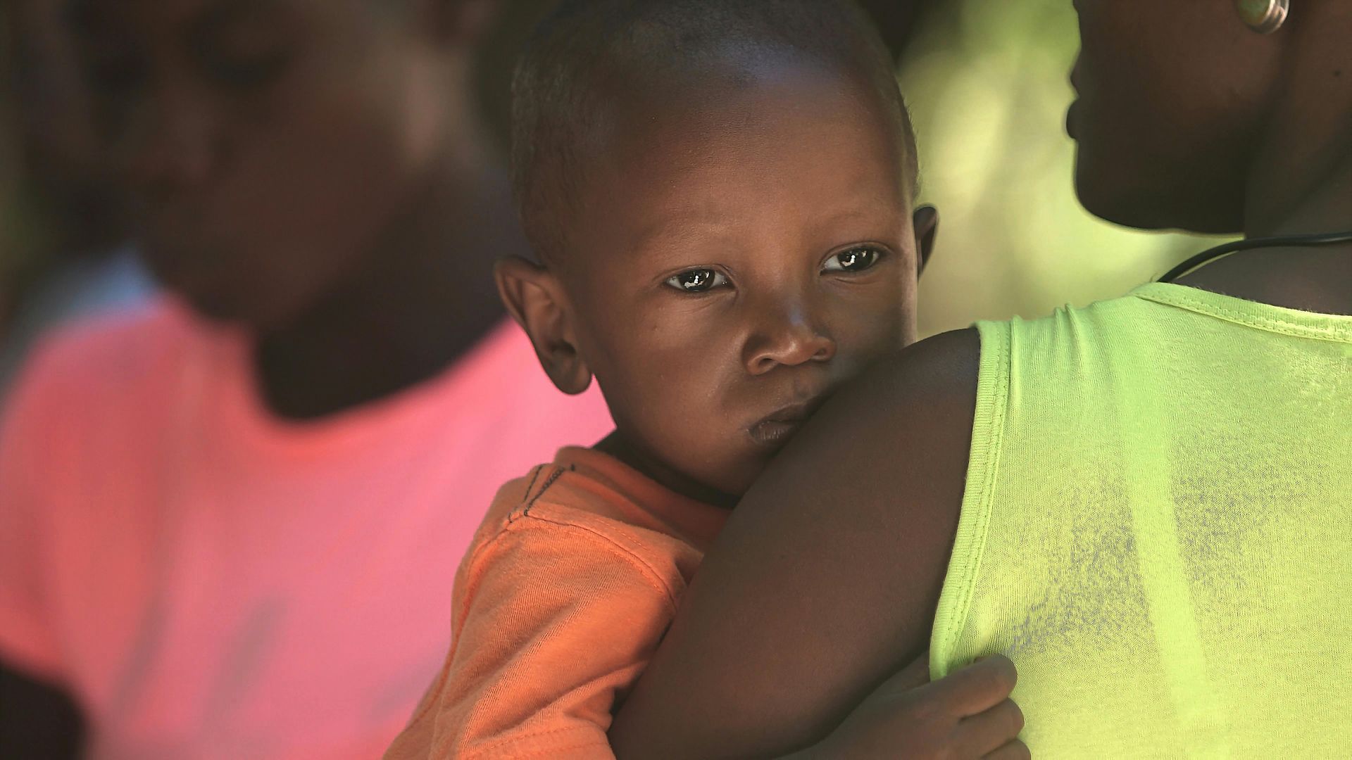A small child wearing an orange shirt is held by a person in a bright yellow top, with another person in pink in the back.