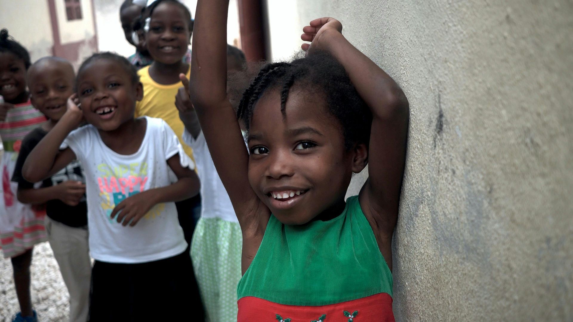 A group of smiling children stand together outdoors, with one child in the foreground wearing a green and red top.