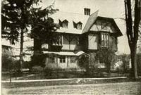 Black-and-white photo of a large Victorian-style house with a steep roof and trees in front