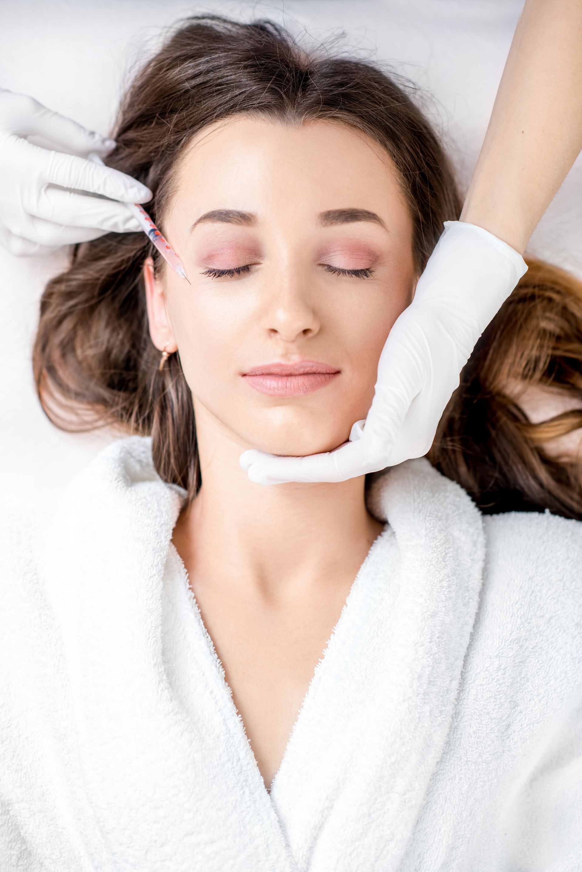A gloved hand injects the forehead of a relaxed woman lying with closed eyes in a soft white robe. A gloved hand injects the forehead of a relaxed woman lying with closed eyes in a soft white robe.