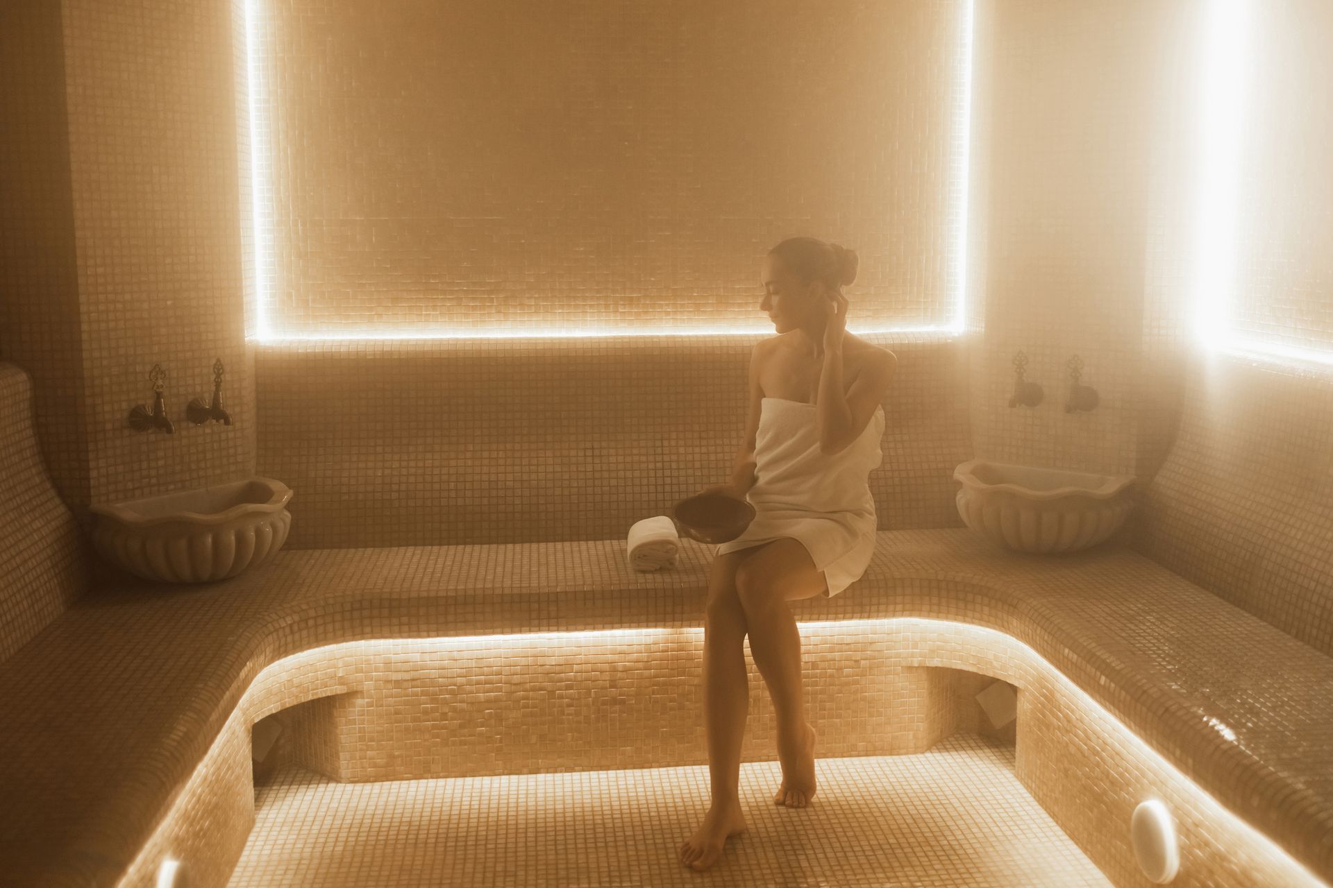 Woman wrapped in a towel, sitting in a steamy, tiled spa room, near basins and glowing lights.