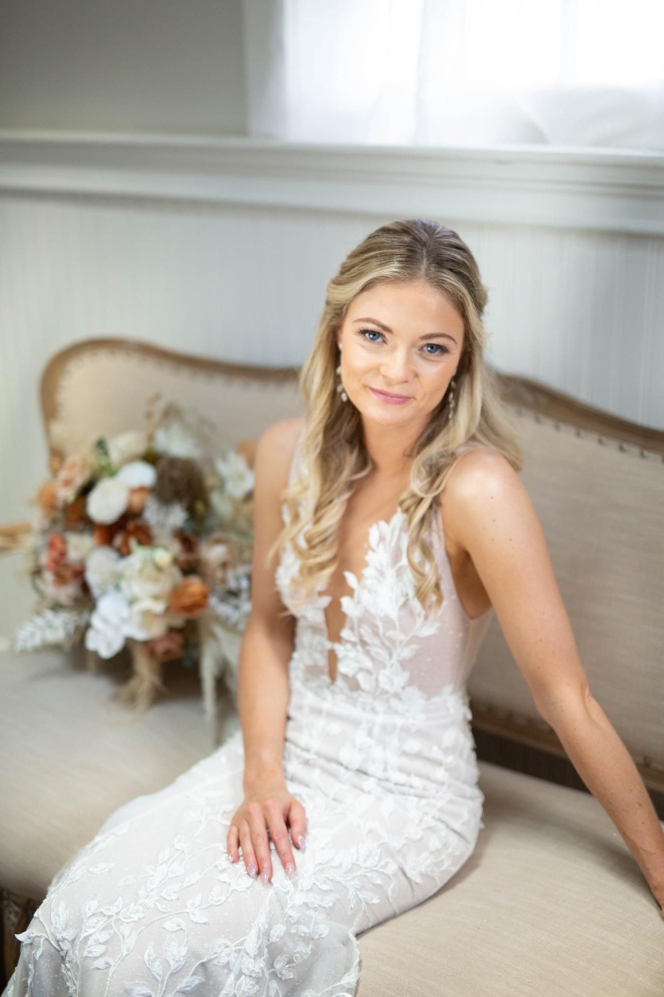 Bride in white wedding dress sits on a beige couch, holding bouquet.