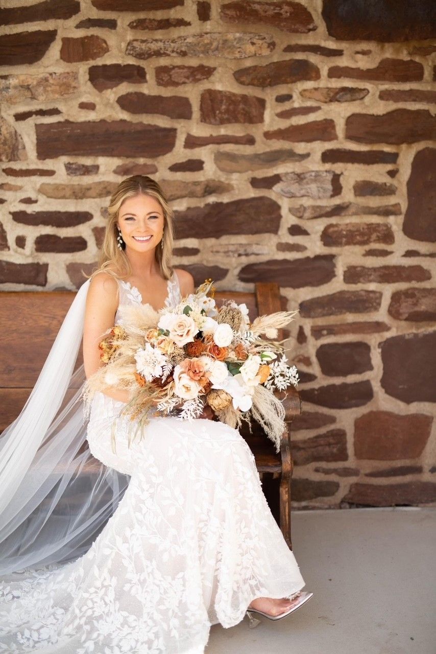 Bride in a lace gown, holding bouquet, seated on a wooden bench in front of a stone wall.