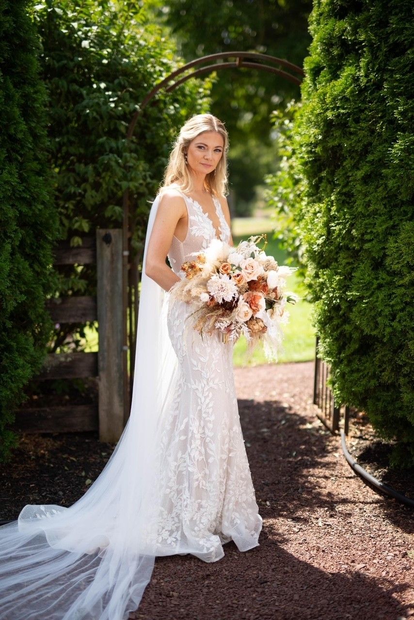 Bride in a sparkling, sleeveless wedding dress holds flowers, standing near an archway with greenery.
