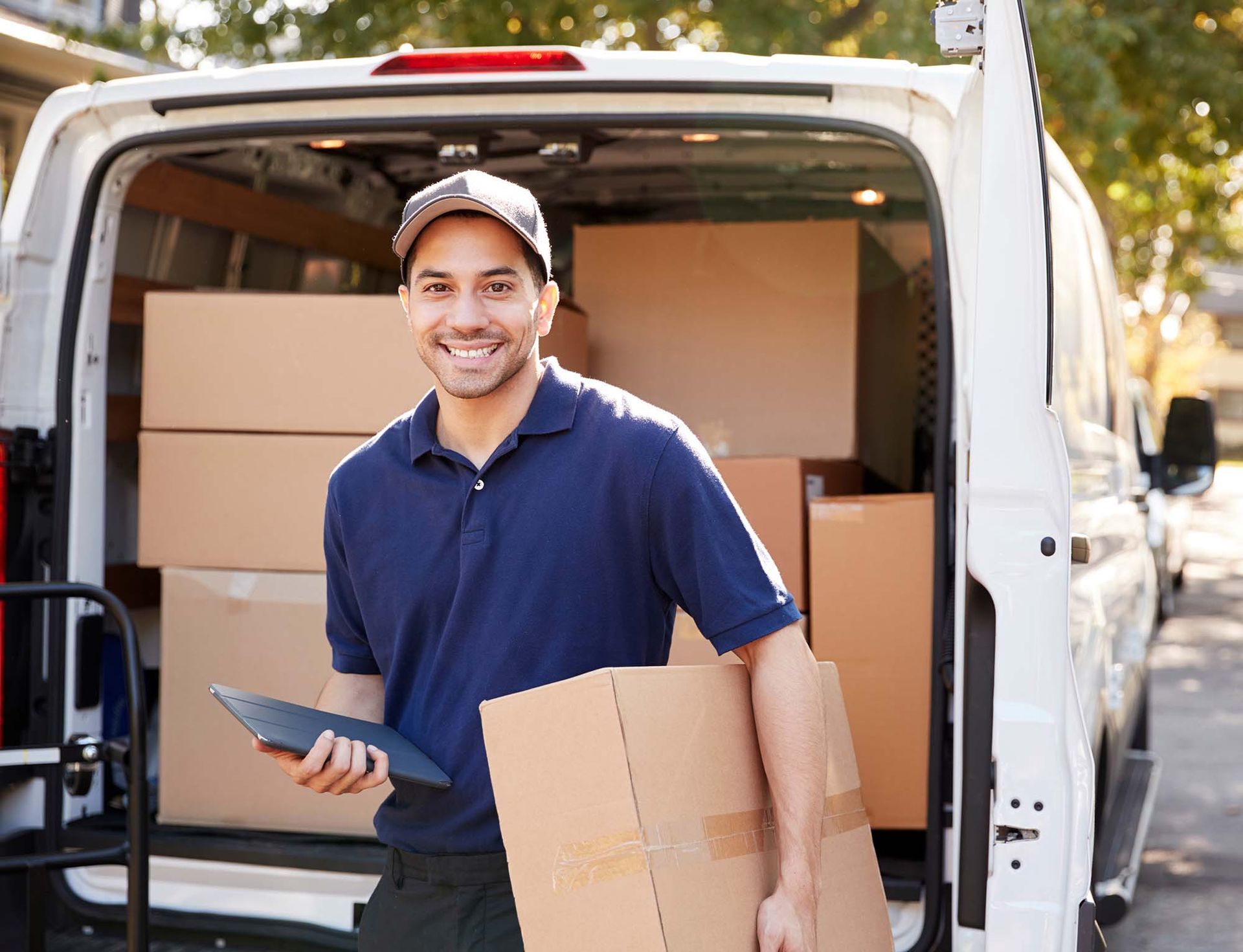 Delivery worker smiling by open van, holding a package and tablet among stacked boxes.