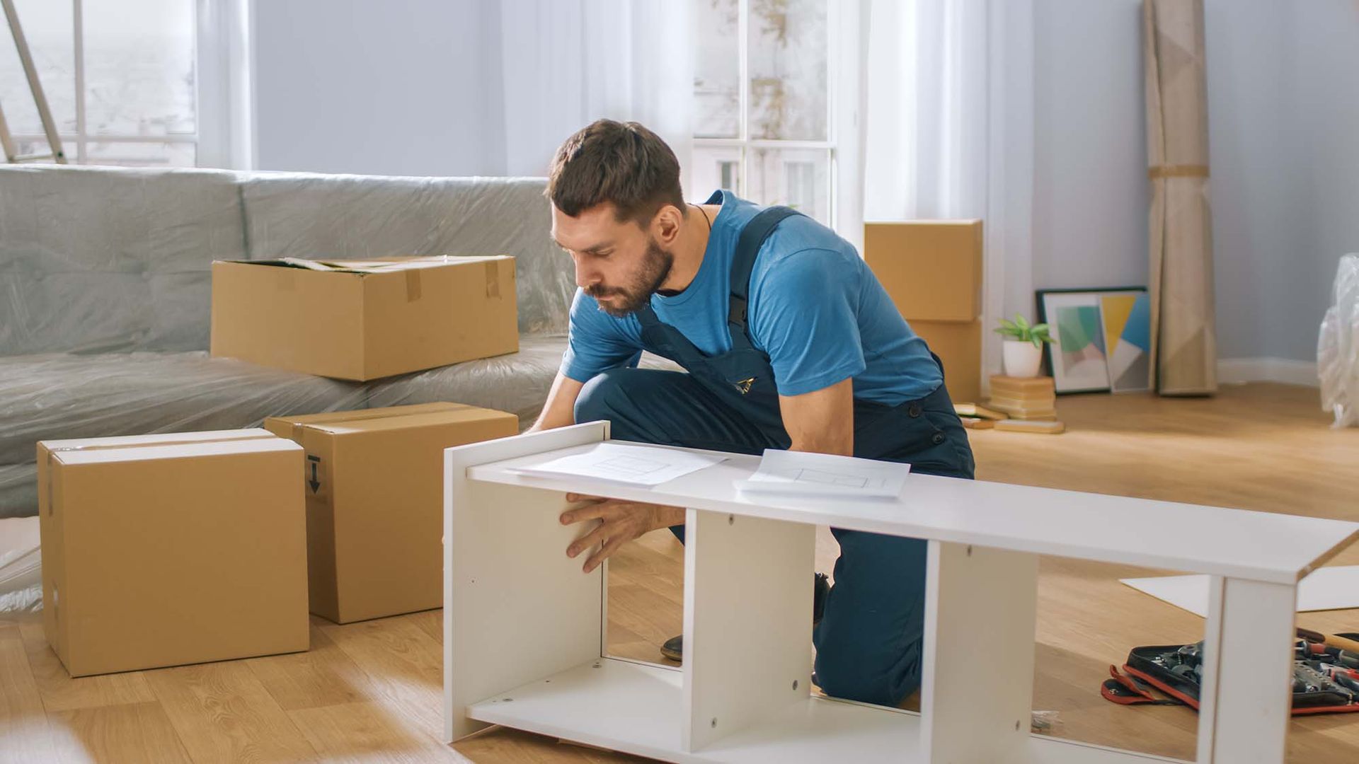 Man assembling a white shelf in a bright room with moving boxes nearby